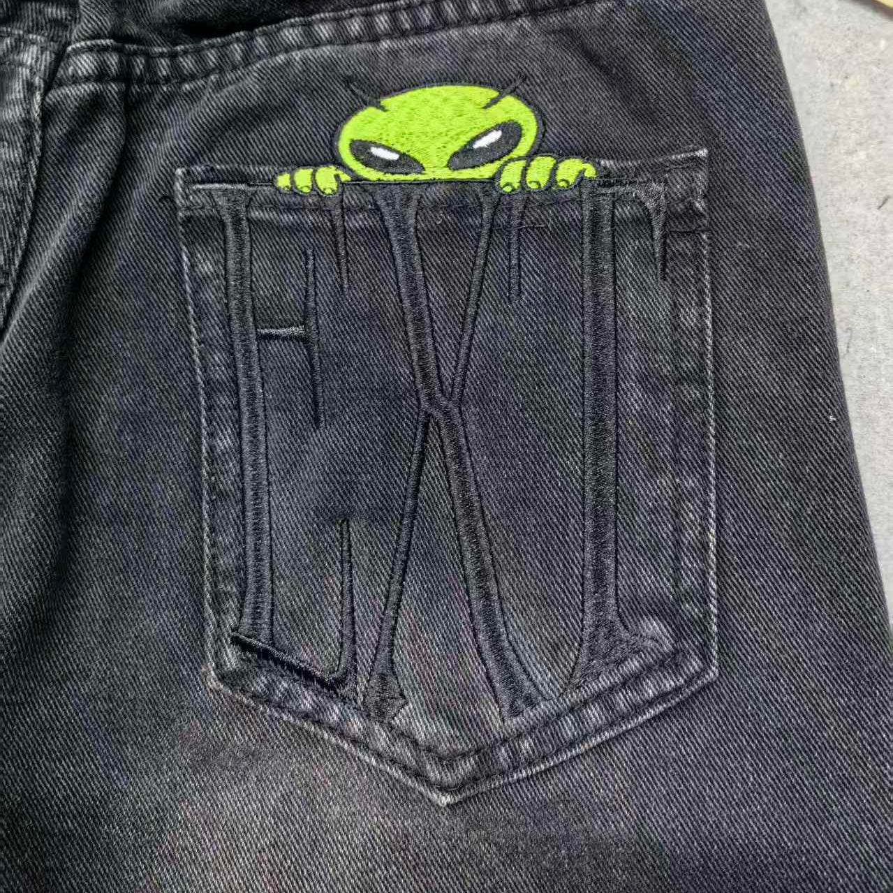 Black denim shorts with a green alien design in the pocket, on a gray surface.